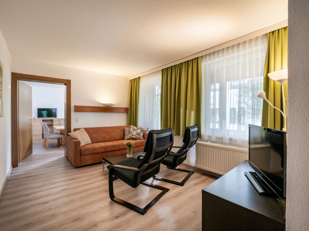 Modernes Hotelzimmer mit Sitzecke, Fernseher und einem Schreibtisch, der im Nebenraum sichtbar ist.