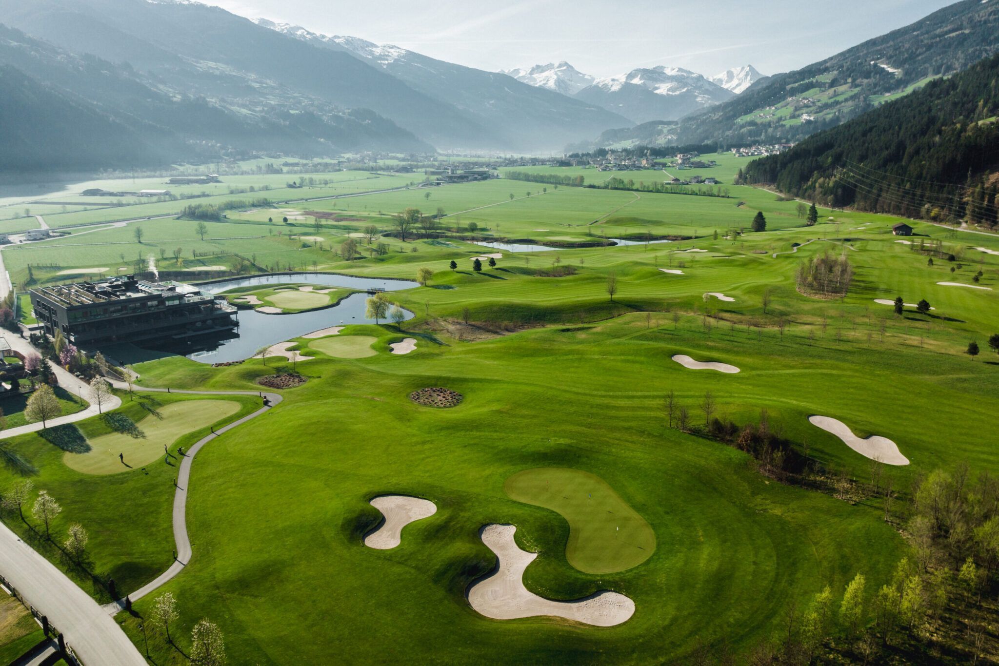 Luftaufnahme eines Golfplatzes mit Grüns, Sandbunkern, einem Wasserhindernis und einem Clubhaus vor der Kulisse üppiger Täler und schneebedeckter Berge.