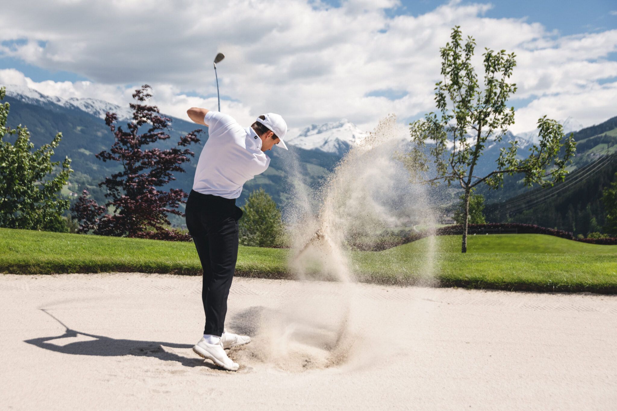 Ein Golfer schlägt in einen Sandbunker und lässt dabei eine Sandwolke aufwirbeln. Schneebedeckte Berge und grüne Bäume bilden die malerische Kulisse unter einem teilweise bewölkten Himmel.