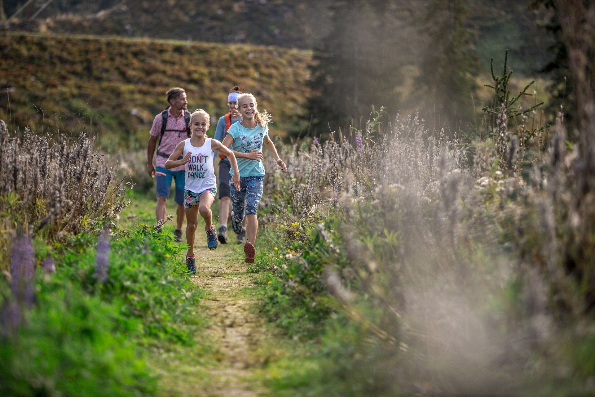 Zwei Kinder laufen einen Pfad entlang, zwei Erwachsene folgen ihnen auf einer Rasenfläche im Freien. Die Landschaft ist von üppigem Grün und hohen Pflanzen geprägt.