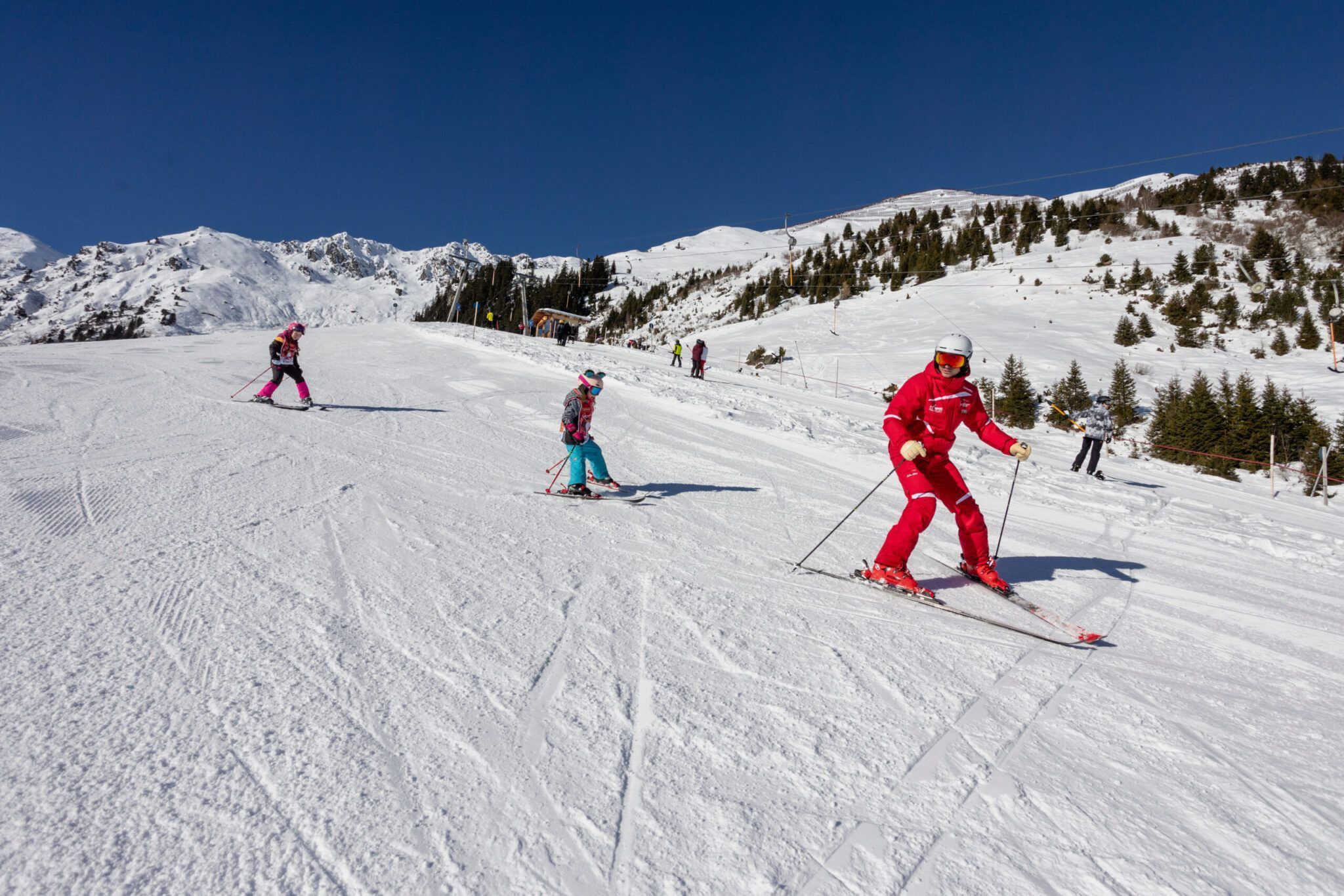 An einem klaren Tag fahren mehrere Skifahrer einen schneebedeckten Berghang hinunter. Eine Person in einem roten Outfit führt eine Gruppe an. Im Hintergrund sind schneebedeckte Berge und Bäume zu sehen.