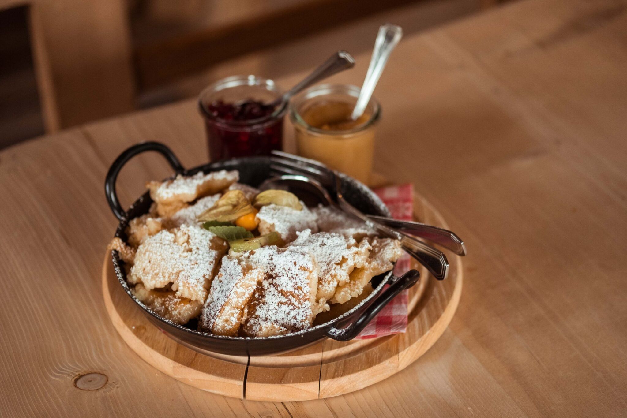 Eine Bratpfanne auf einem Holztablett enthält mit Puderzucker bestreute Dessertstücke, dazu zwei Löffel. Kleine Gläser mit roter und gelber Marmelade sowie Löffel stehen neben der Bratpfanne.