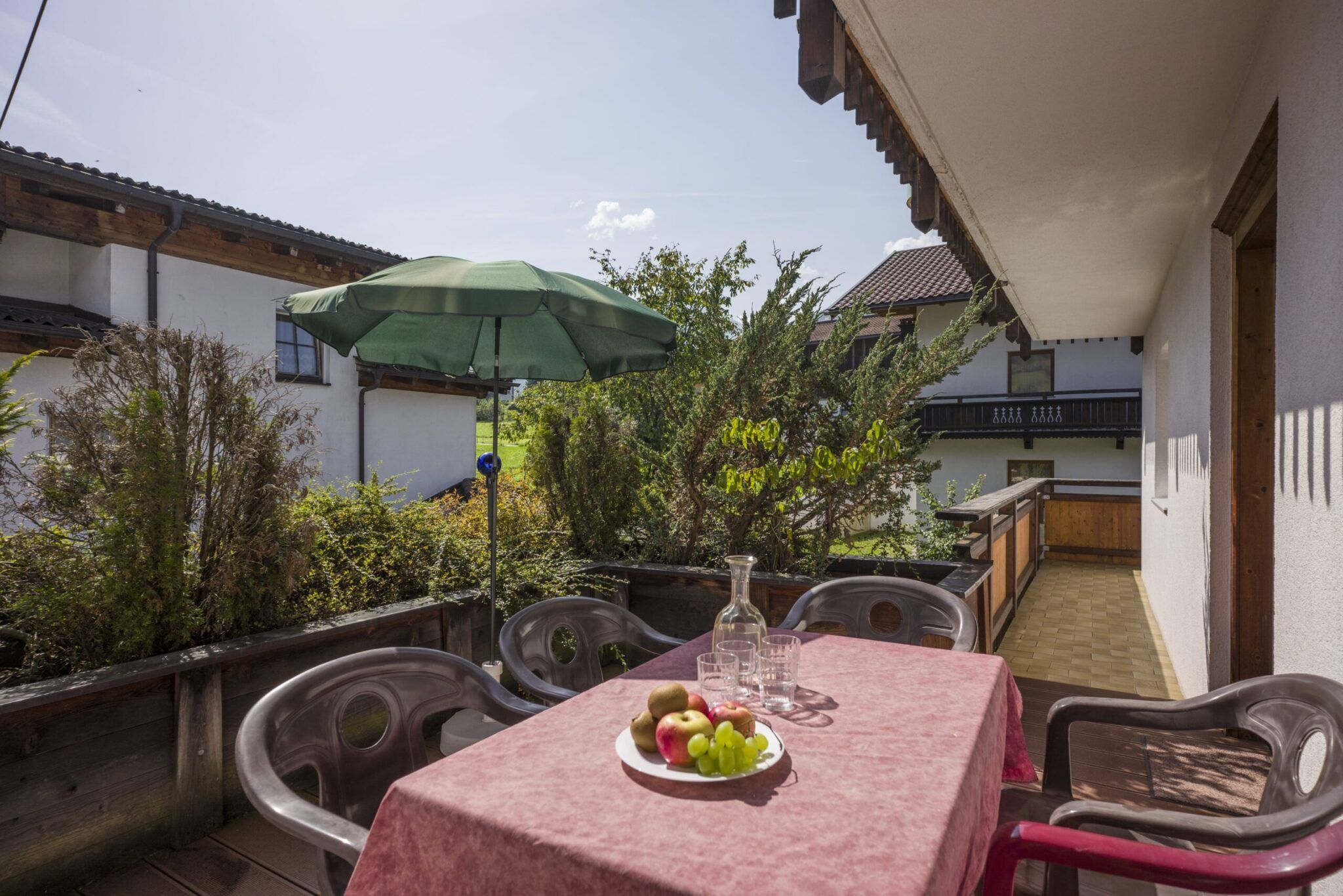 Buitenterras met een tafel bedekt met een rood tafelkleed, een fruitschaal en een groene parasol, omgeven door groen.