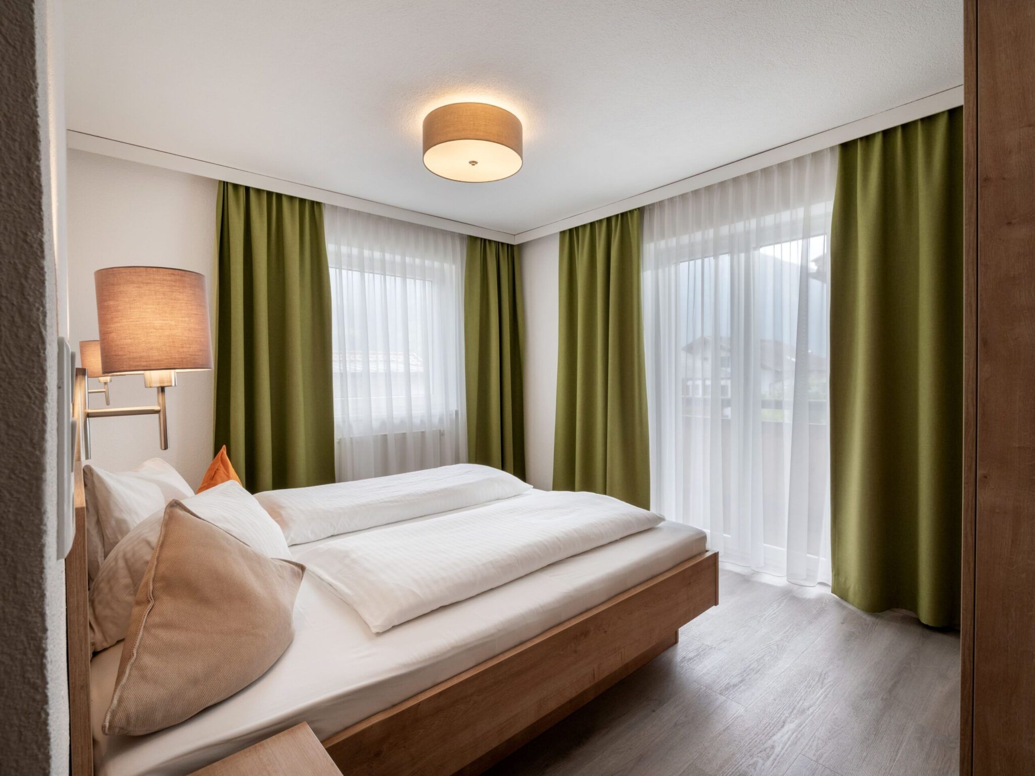 Een gezellige slaapkamer met een tweepersoonsbed, wit beddengoed, groene gordijnen, houten meubels en een plafondlamp.