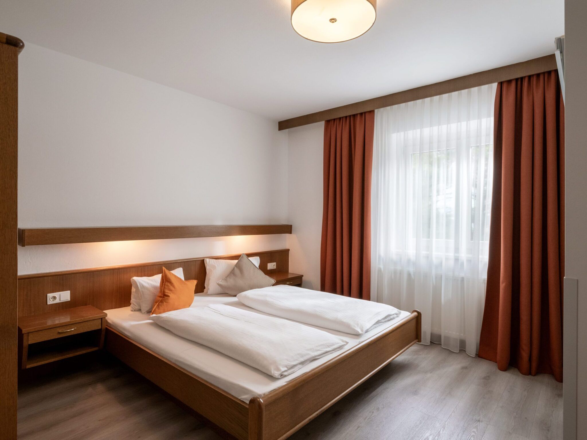 Een nette hotelkamer met een tweepersoonsbed, twee nachtkastjes en een raam met witte en rode gordijnen.